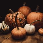 A photograph still life of 8 different pumpkins of different sizes. Some are orange, some are white. The lighting is dramatic. The pumpkins are sitting on a wooden floor and the background is black.